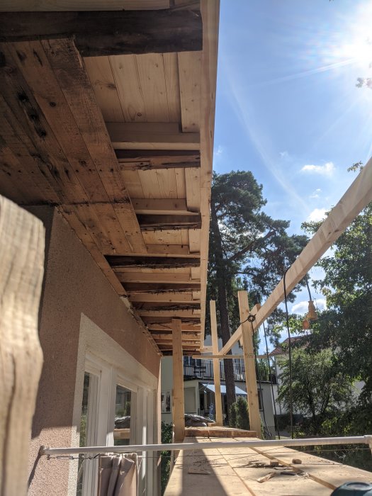 Renoverad balkong i halvfärdigt skede med ny råspont och hemmasnickrad byggställning mot soligt landskap.