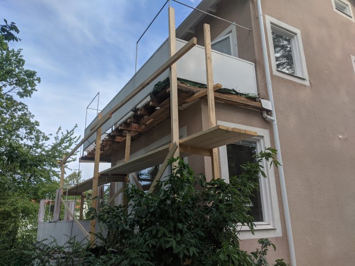 Pågående renovering av en balkong med nytt träbjälklag och byggställning synlig, intill en ljusbrun husfasad.