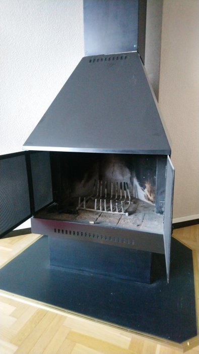 Svart braskamin med öppna dörrar som visar eldstaden inuti, placerad på ett parkettgolv.