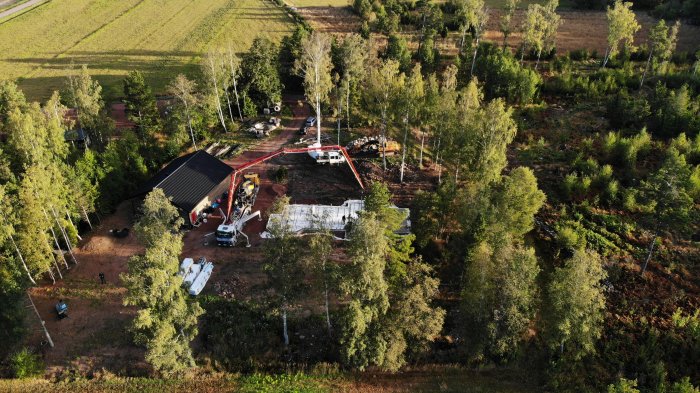 Ovanifrån vy av byggplats med gjutningsarbete för husgrund omgiven av träd och fält.