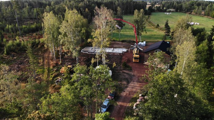 Drönarbild på husgrundsgjutning med betongbil och gjutarm, omgiven av grönska och skog.