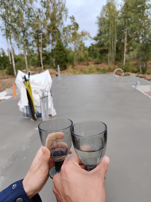 Två personer skålar med läsk på nyligen gjuten betongplatta, omgivna av träd och byggmaterial.