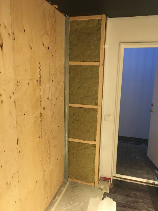 Nybyggd vägglåda med isolering i träreglar och en del plywood uppsatt nära en dörröppning.