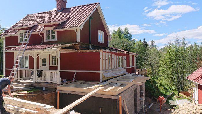 Byggplats med stomresning vid rött hus, träreglar på grund och delar av en demonterad veranda syns.