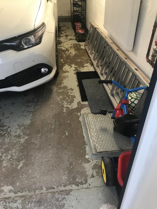 En vit bil i ett garage med slitna och fläckiga betonggolv, leksaksbil och föremål på sidan.