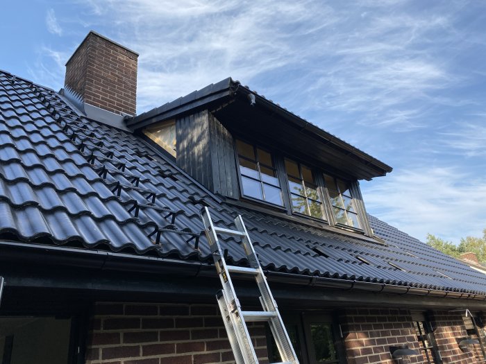 Svart takpannor på ett hus med tegelväggar, stege lutad mot huset, skorsten och vindfönster mot en himmel med cirrusmoln.