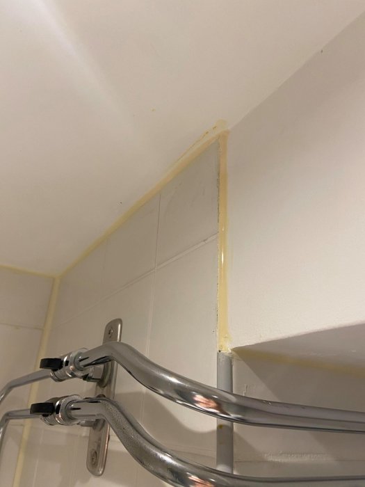 Gulaktig, utsmetad fog längs taket och vit kaklad vägg med duschkran och rör i förgrunden.