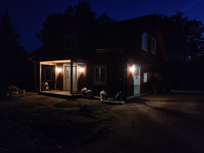 Tvåvåningshus med tända utelampor i skymningen, byggmaterial framför huset.