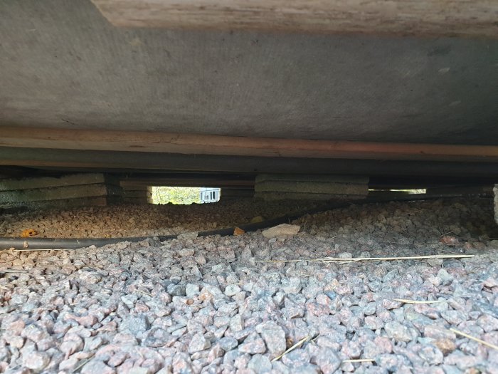 Vy under en friggebod som visar golvets understruktion med hardboard och plankor ovanför grus.