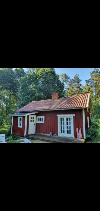 Rödmålad timmerstuga från 1850 med vita knutar och fönster, omgiven av grönska.