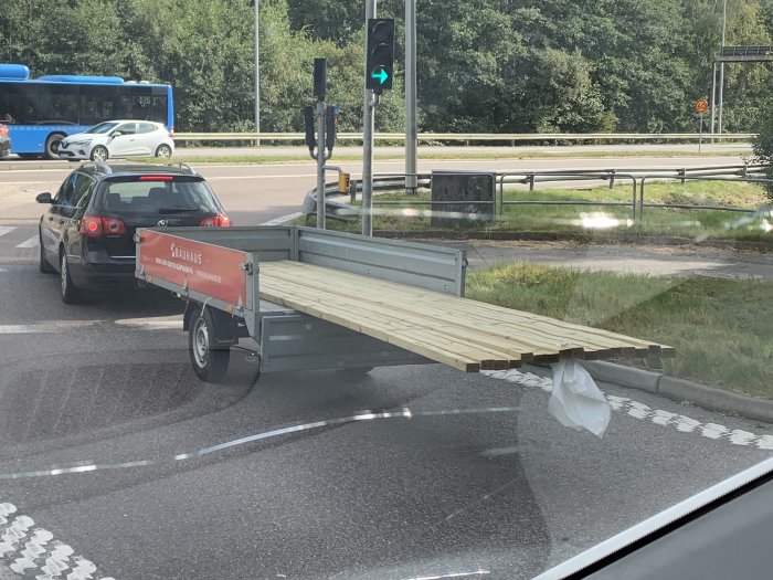 Bil med släpvagn överlastad med långa plankor som sticker ut bakåt vid trafikljus.