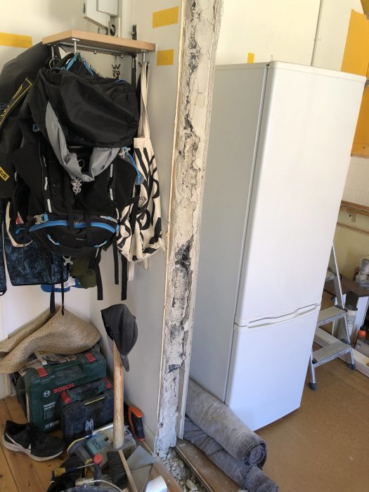 Exposed concrete wall edge next to a white fridge, surrounded by construction tools and materials.