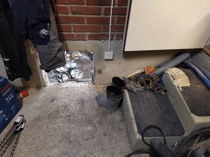 En rörig garagehörna med murbruksspill, verktyg, arbetshandskar och armeringsnät vid en tröskel.