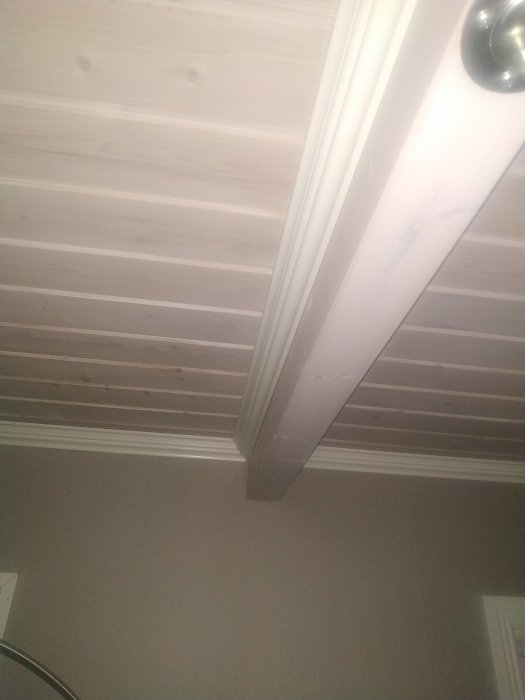 Vy snett uppåt mot en takbjälke i linje med väggen, med vitmålad träpanel i taket.