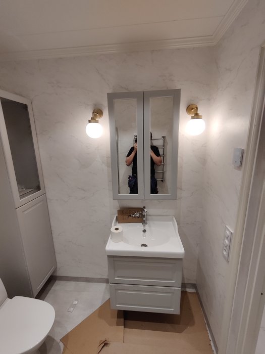 Nyrenoverad toalett med vit inredning, marmormönstrade väggar och guld detaljer på lampor.