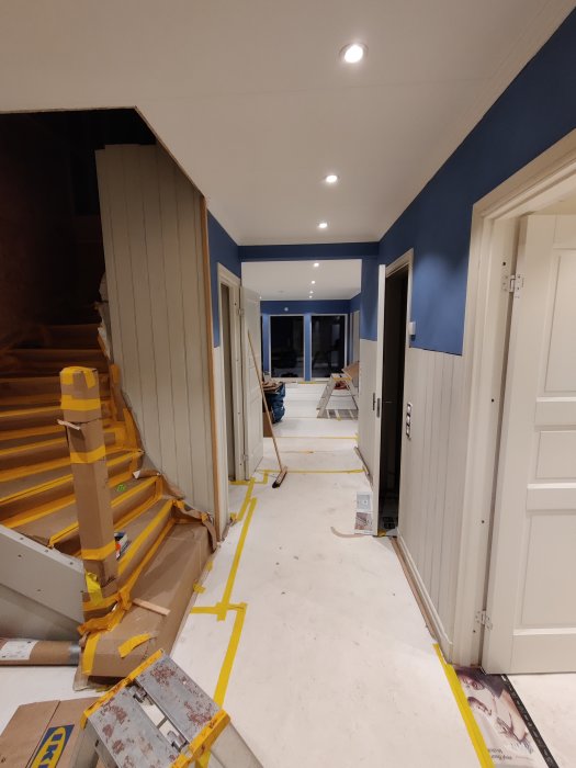 Renoveringsarbete i en hall med blå väggdekoration, skyddstejp på golv och trapp, samt installerade spotlights.