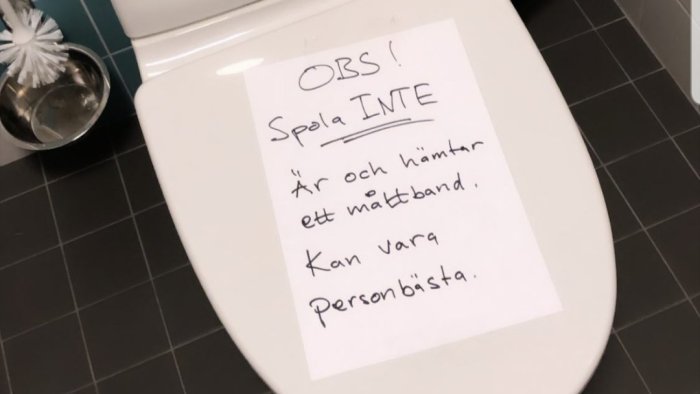 Lapp med text "OBS! Spola INTE 'är och hämtar ett måttband, Kan vara personbästa.'" på en toalettsits.