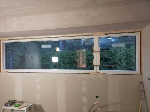 Nymålad innervägg med spacklade skarvar och nyinstallerat panoramafönster som visar trädgårdsutsikt.