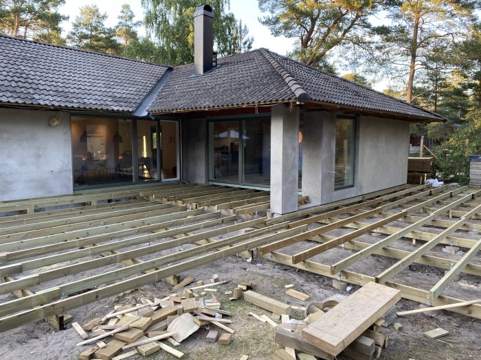 Påbörjat trädäck med längsgående reglar och dolda skruvar vid ett hus, träbitar och verktyg på marken.