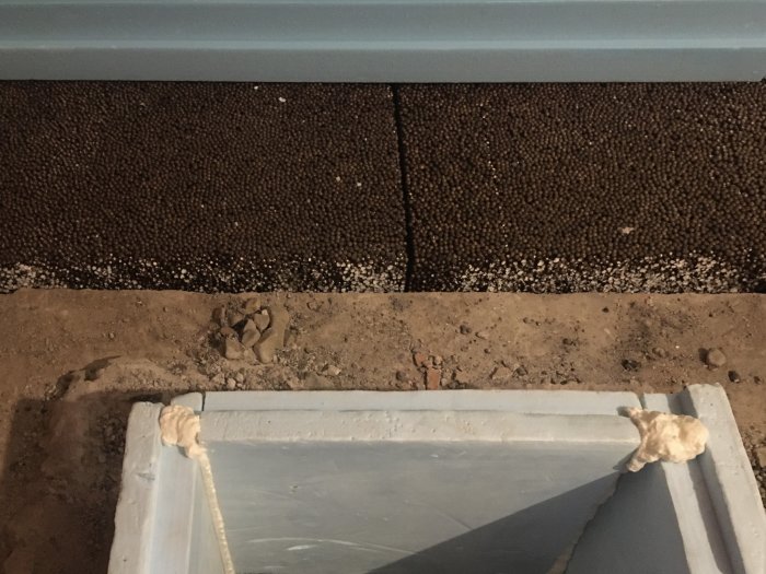 Inspektionslucka i källargolv med grå ram, intill dränskivor och murbrock.