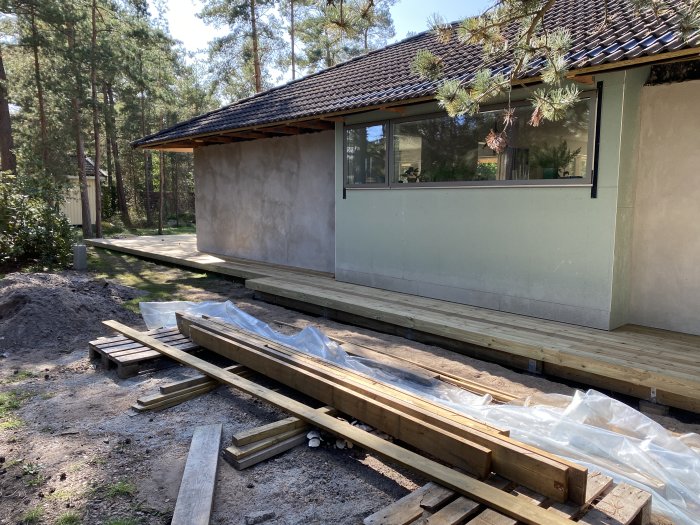 Nybyggd trätrall runt hus med byggmaterial och halvfärdig fasad i skogsmiljö.