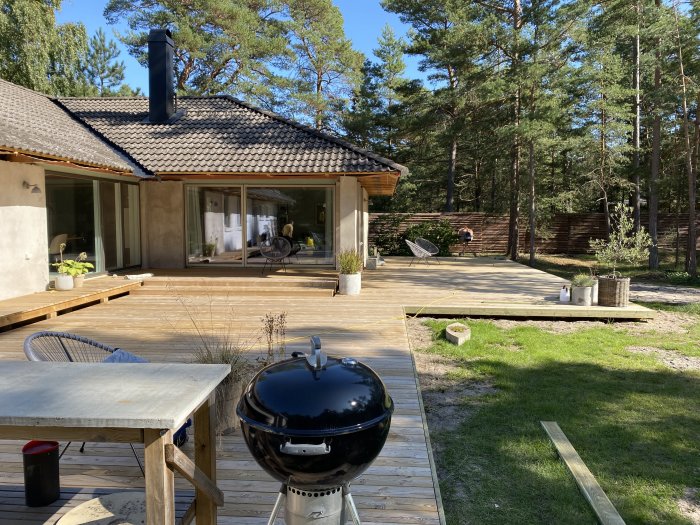 Nybyggt trädäck vid hus, nästan klart, med trädgårdsmöbler och grill, omgivet av träd och en gräsmatta.