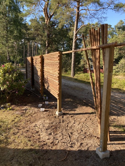 Delvis färdigställd rundvirkesstaket med 900 träpinnar i en skogsmiljö, med ytterligare pinnar som väntar på installation.