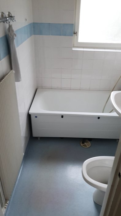 Äldre badrum i behov av renovering med vit och blå kakel, badkar, toalett och golv som ska bytas ut.