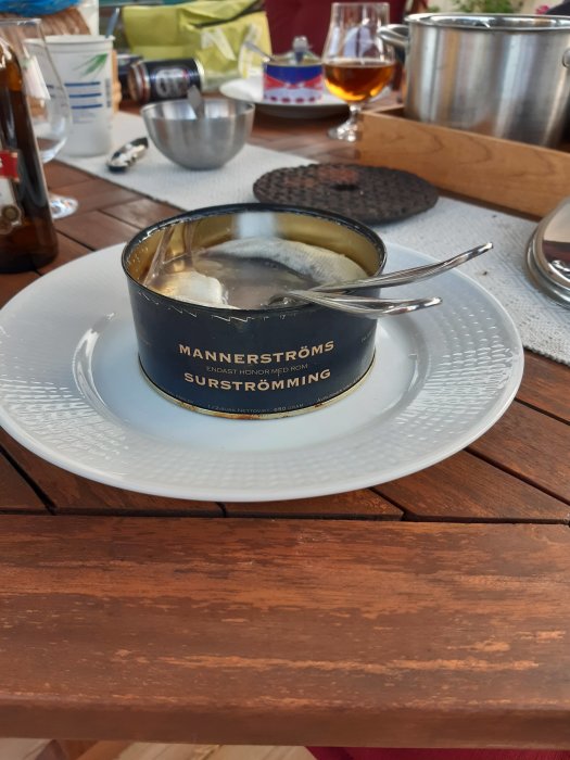En öppnad burk med Mannerströms surströmming på en vit tallrik vid ett middagsbord med drycker och köksutrustning i bakgrunden.
