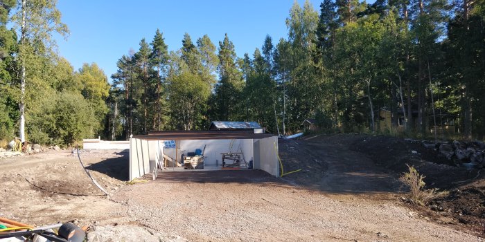 Husbygge på grundnivå med exponerad carport och källare, omgiven av en skogsmiljö och klar himmel.