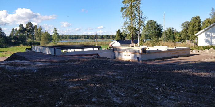 Byggplats med grunden för huset, stålkonstruktioner och suterrängvägg synlig i ett landskap med himmel och träd.