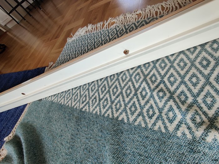 Äldre vit dörrkarm ovanpå två mattor med synliga skruvhål och en borttagen list på ett trägolv.