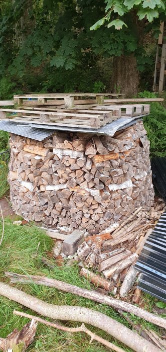 En oregelbunden vedstapel med barken åt olika håll, täckt av en presenning och träpallar, belägen utomhus.