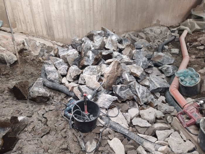 En byggarbetsplats med spräckta stenblock, en bergborr och slangar vid en betongvägg, redo för ytterligare arbete.