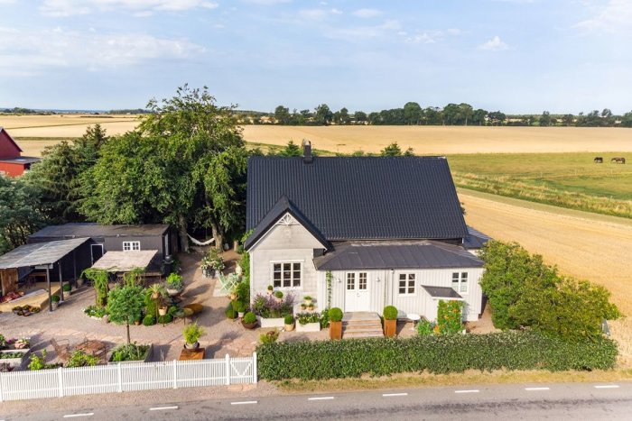 Ett traditionellt svenskt hus med vita fasader och svart tak, inramat av grönska med åkrar i bakgrunden.