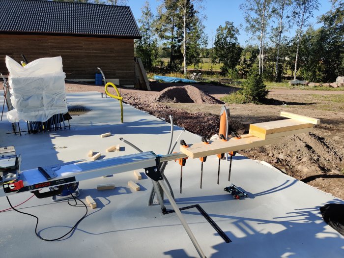 Virkeskapning för husbygge med sågbord och timmer på en nyligen gjuten betongplatta utomhus.