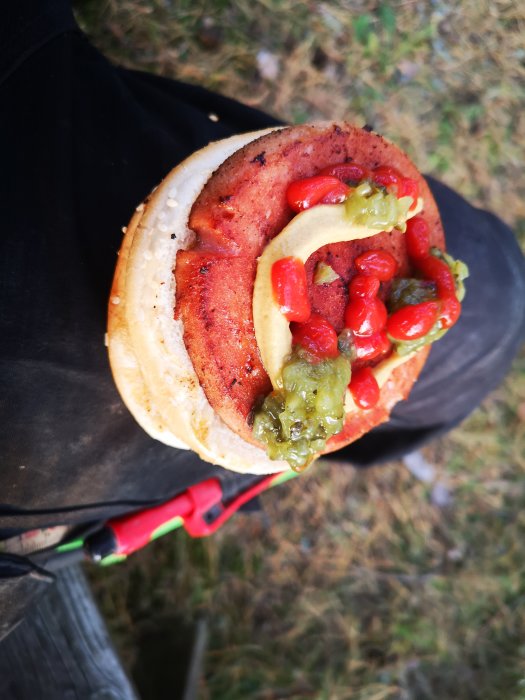 Grillad parisare i bröd med senap och ketchup, hållen i hand utomhus.