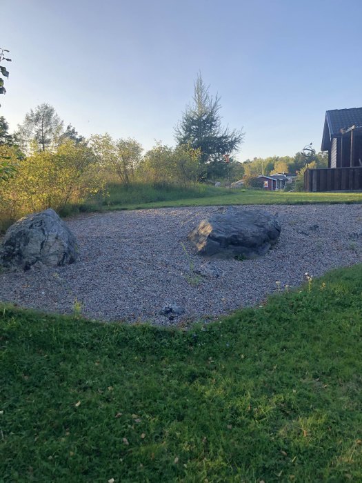 Grusparti på en tomt med stora stenar omgiven av gräs och träd bakom, bredvid en byggnad.