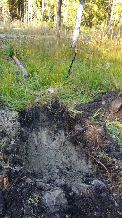 Testgrop grävd på en tomt med sand och lera, omgiven av grönt gräs och skog i bakgrunden.