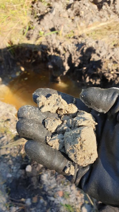 Hand med handske håller sand- och lera-prover med en grävd grop fylld med vatten i bakgrunden.