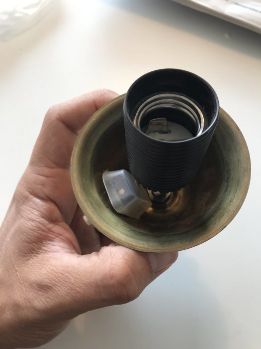 Hand håller en isärplockad lampsockel med synlig lamphållare och tryckbrytare i mässing.