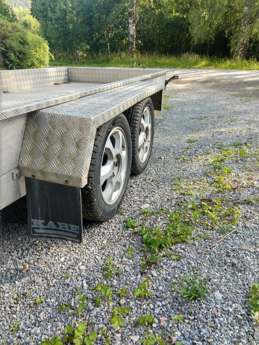 Del av en ombyggd släpvagn med märkning 'KABE', rutmönstrad metall och dubbla hjul, parkerad på grus.