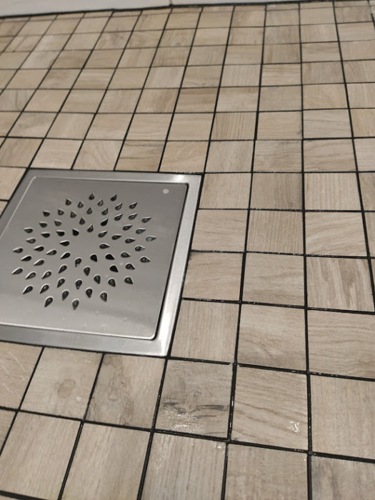 Renoverat badrumsgolv med mosaikklinker och en golvbrunn, ojämn nivå nära brunnen.