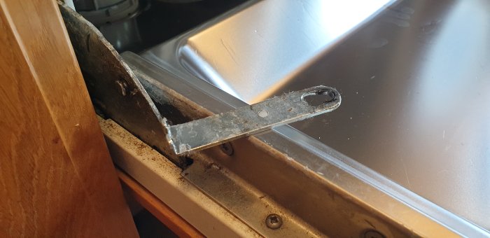 Metallfäste med hål och slitage under diskmaskin, nära träpanel och diskbänk.