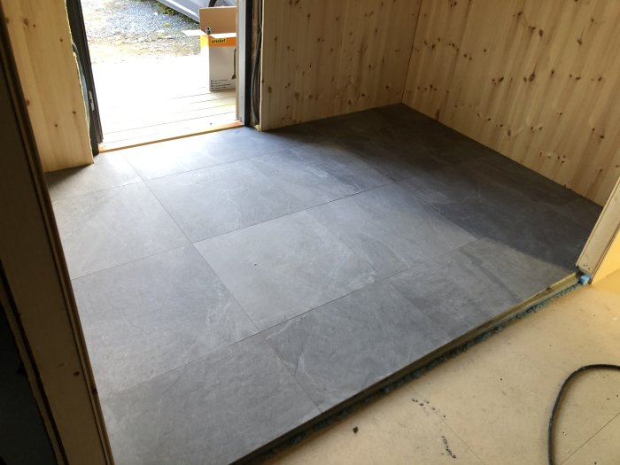 Nyklätt golv med stora 60x60 cm plattor i en öppen hall mot en ytterdörr, omgiven av träväggar.