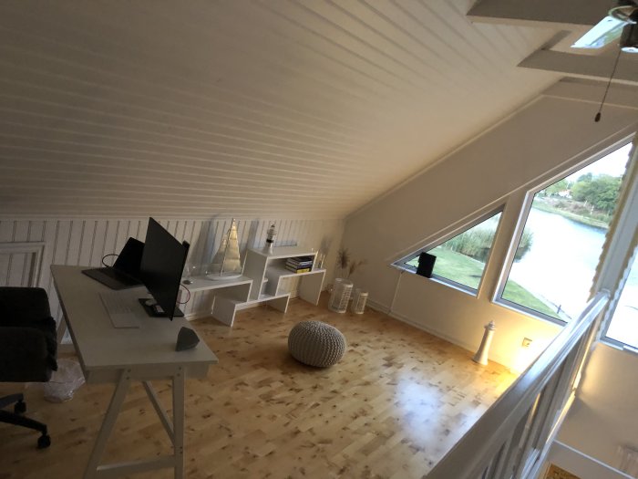 Renoverad övervåning med ny arbetsplats, vita hyllor och utsikt över vatten genom fönster.