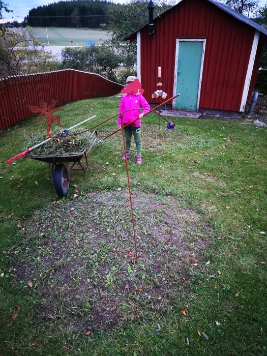 Barn i rosa jacka puttar en skottkärra med trädgårdsavfall framför röd stuga och gräsmatta.