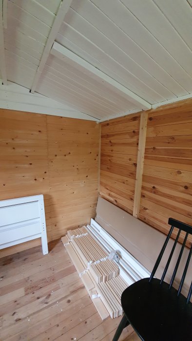 Innerutrymme av en bod med träpanelväggar, oisolerat tak och trägolv med isoleringsmaterial på golvet.