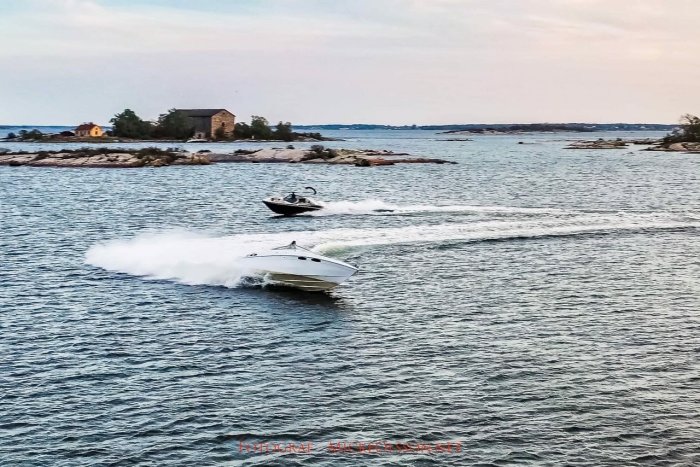Båtar kör i formation i skärgården med ö och stenhus i bakgrunden.