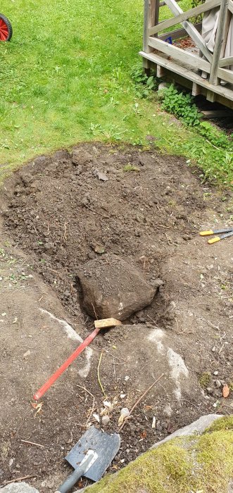 Grävd mark med stenar och rötter, en spade och en slägga, vid grunden till en trappa i en trädgård.